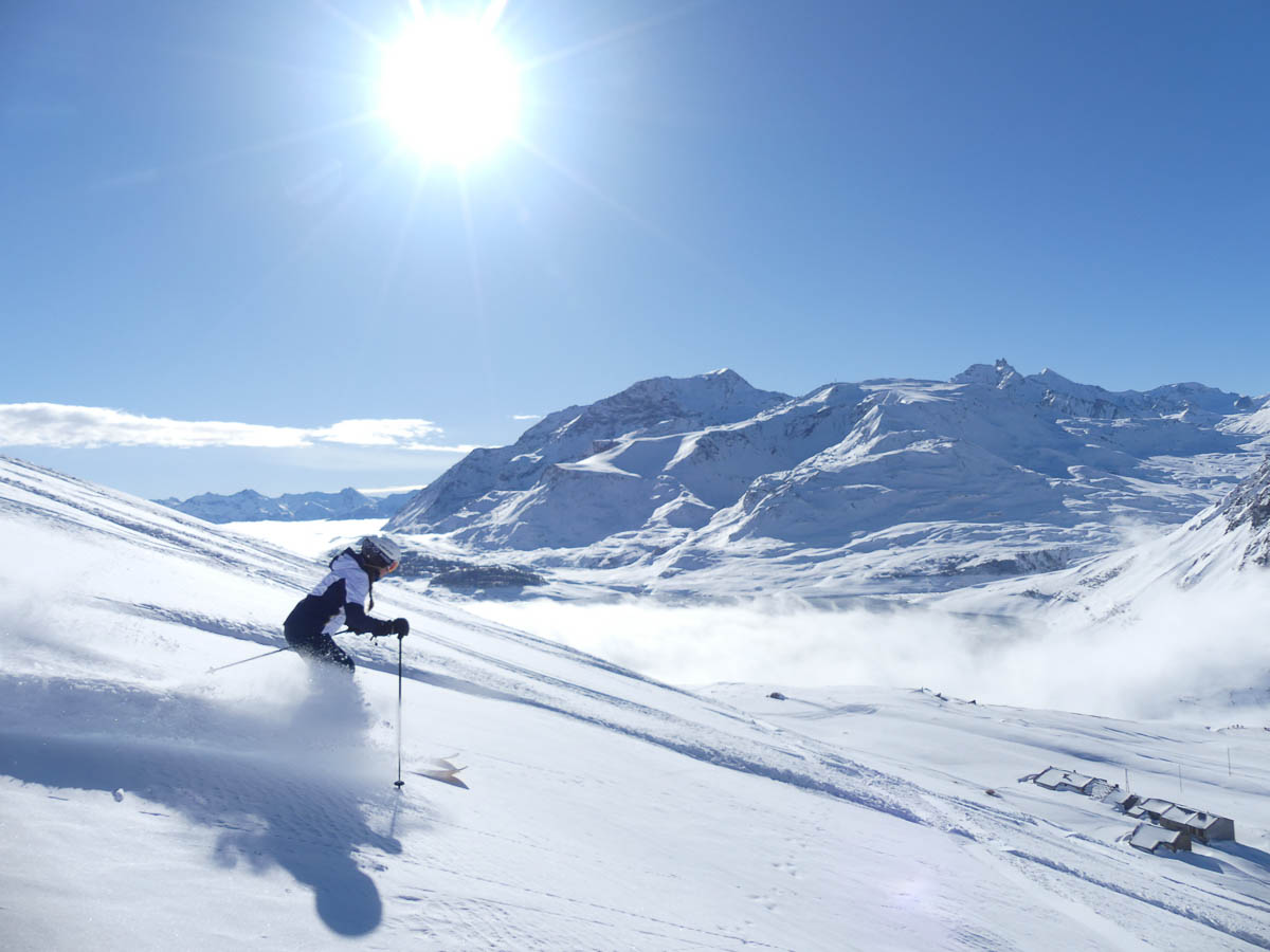 solo skier on mellow off piste, grand mountains and sun shining behind them as they descend