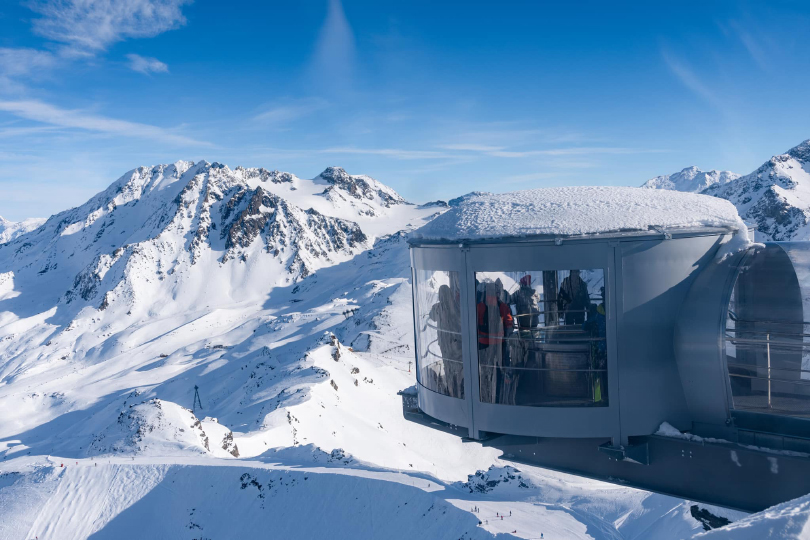 a viewing room juts out over a mountain scene, skiers on a cat track far below