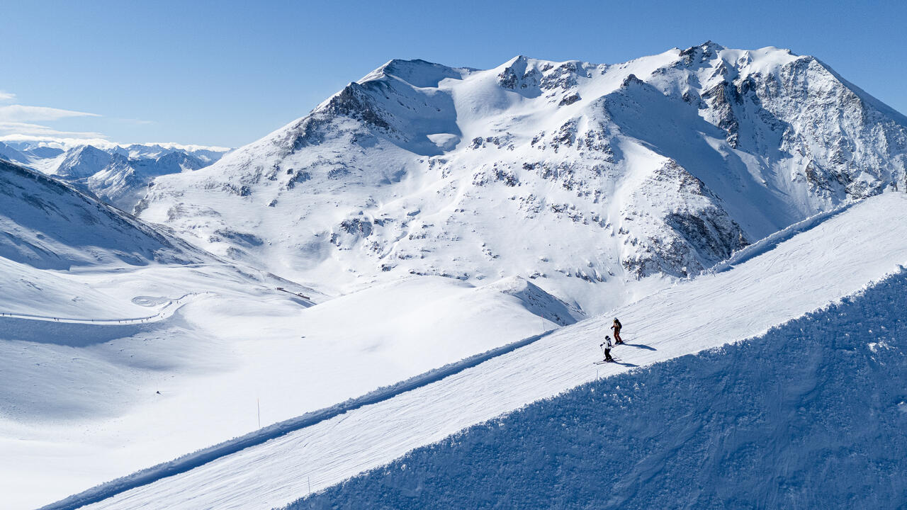 two skiers on a skinny ribbon of piste, steep faces to each side, high in the snowy mountains on a blue-sky day. A piste with more ant-sized skiers winds in the distance