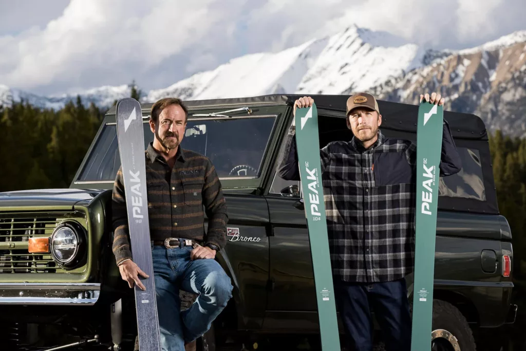 Bode Miller holds up two skis from his brand Peak, leaning against a Landrover in the mountains. Another man holding up a pair next to him
