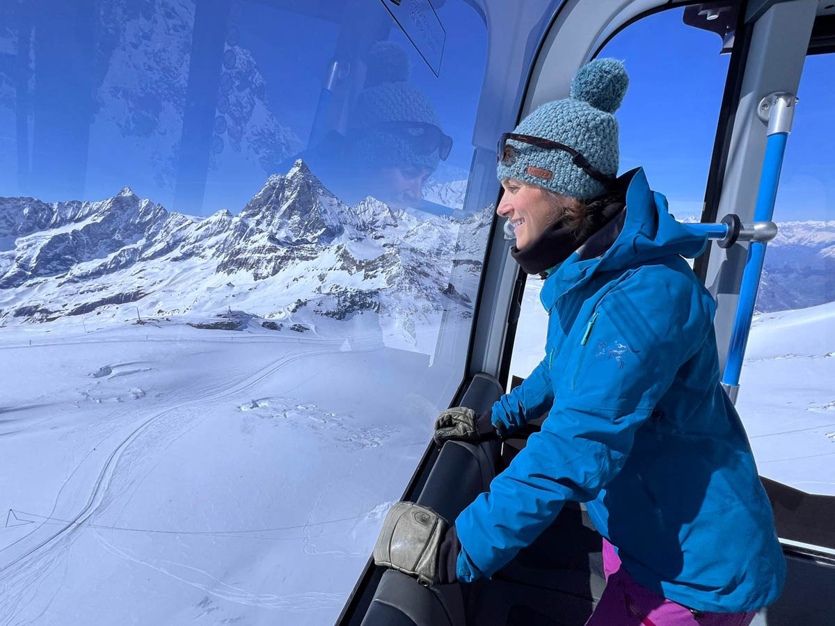Welove2ski editor Nicola wearing blue stands inside a gondola looking out at the view below - a view of high snowy mountains and ski-tracked snow beneath